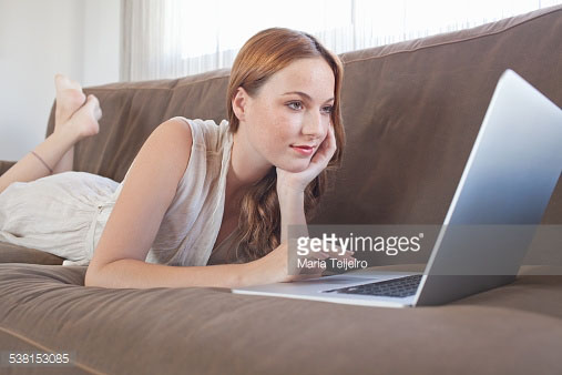 Bijschrift:Attractive young woman lounging and relaxing on a sofa at home, using a laptop computer. Indoors lifestyle